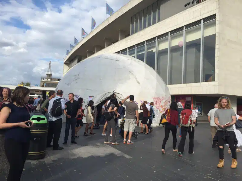 A group of people gather outside a large white dome-shaped structure near a modern building with glass windows and flags on the roof, under a partly cloudy sky. Some people are talking, walking, or taking photos.
