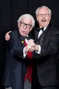 Two older men wearing dinner jackets stand close together, holding hands as if dancing. Both have surprised or joyful expressions on their faces. They are in front of a plain, dark background.