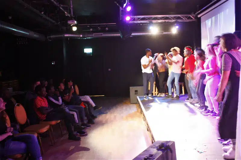 A group of people on stage energetically performing or celebrating, while an audience sits and watches in a dimly lit room with purple stage lights.