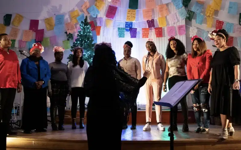 A choir of ten people stands on a stage, singing as a conductor leads them. Behind them are colourful hanging decorations and a lit Christmas tree. The group wears festive clothing and floral headbands.