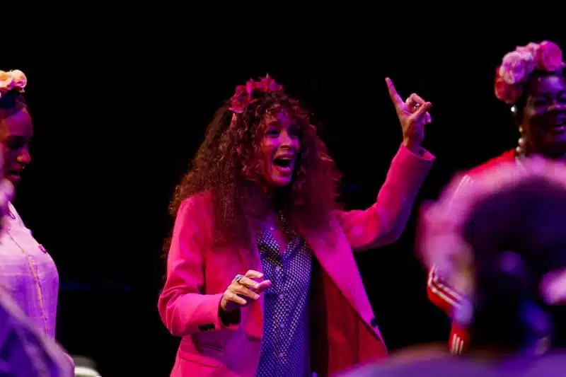A woman with curly hair, wearing a pink blazer and a flower crown, smiles and gestures with her hand raised during a lively event, surrounded by others in festive attire and colourful lighting.