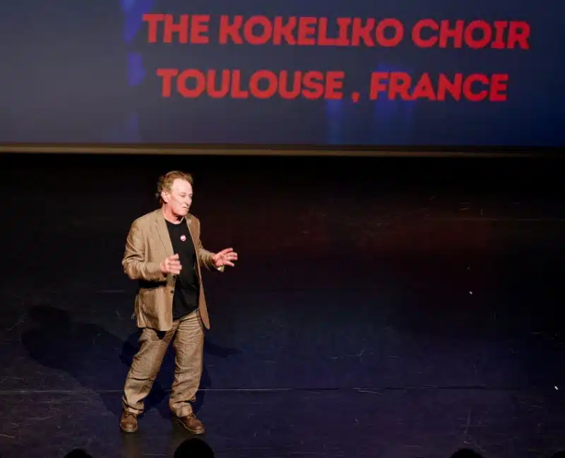 A person in a tan suit speaks on stage with the words THE KOKELIKO CHOIR TOULOUSE, FRANCE projected in red letters on a screen behind them.