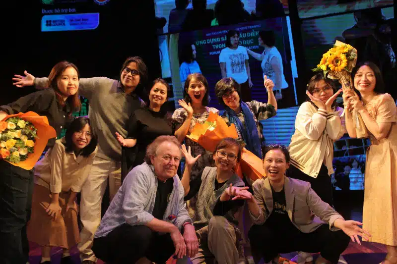 A group of people pose cheerfully on stage, some holding bouquets of flowers. They are smiling, making playful gestures, and standing in front of a large screen displaying images and text.