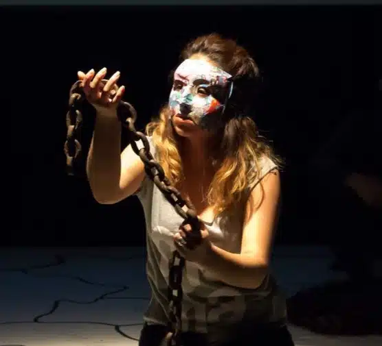 A woman with long hair wearing a colourful artistic mask holds a heavy chain in one hand, reaching forward under dramatic lighting on a dark stage.