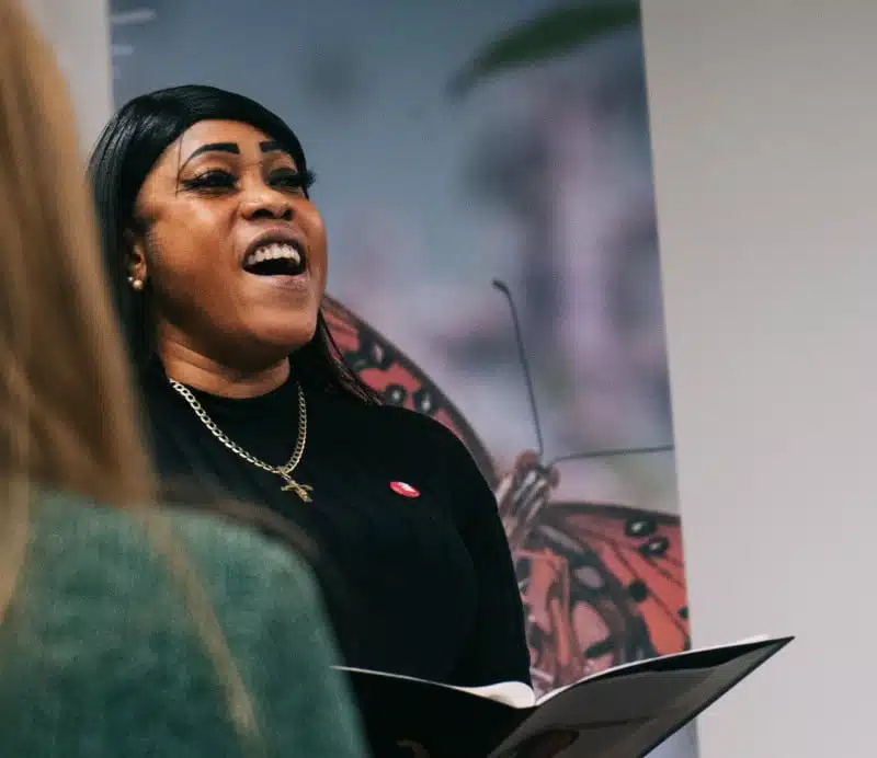 A woman smiles as she sings or speaks, holding an open folder. She wears a black top, gold necklace, and cross pendant, with a butterfly image in the background. Another person is blurred in the foreground.