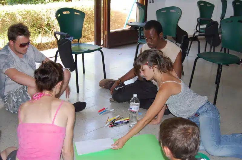 Four young people sit on the floor in a circle indoors, working with markers and paper, appearing to collaborate on an art or group project. Empty green chairs are in the background, and sunlight streams through open doors.