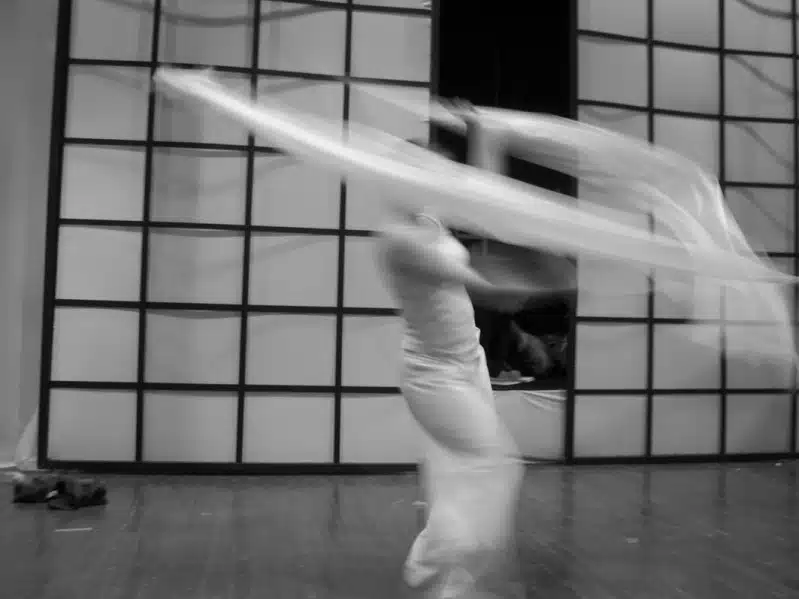 A blurred figure in white dances energetically with long flowing fabric in front of a grid-patterned wall, capturing a sense of motion and grace on a wooden floor.