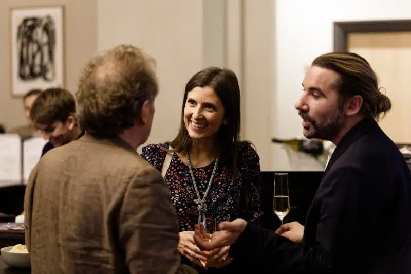 Three people are standing together indoors, engaged in conversation. One woman is smiling and holding a drink, whilst the two men, also holding drinks, are talking. It appears to be a social or networking event.
