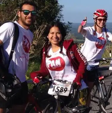 Three people wearing white shirts with red logos are standing outdoors with bicycles. One woman in front is smiling, another in the background is giving a thumbs-up, and a man stands to the left, also smiling.