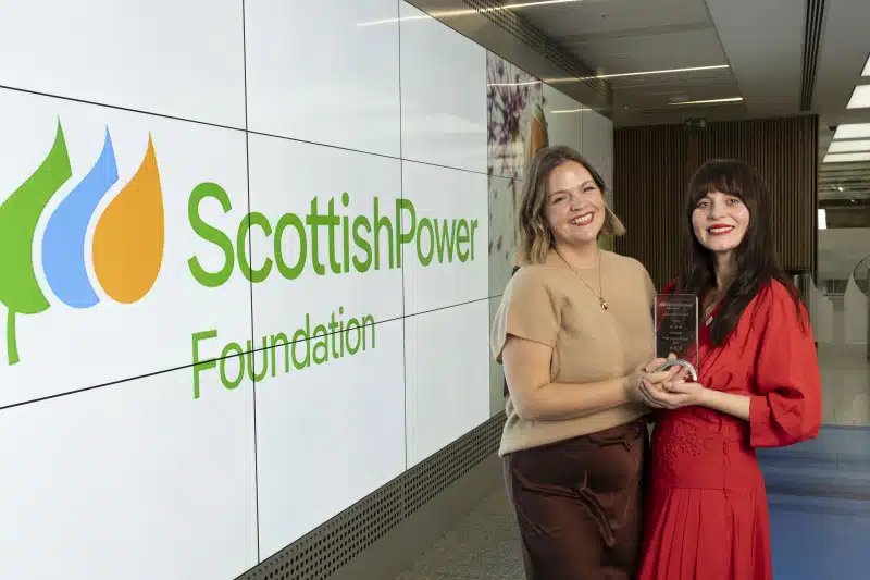 Two women smiling and holding an award stand in front of a large screen displaying the ScottishPower Foundation logo inside a modern building.