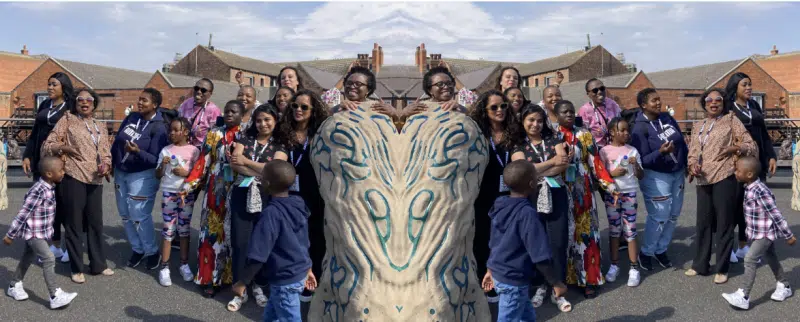 A group of people, including adults and children, stand and pose together outdoors in front of brick buildings. The image has a mirrored effect, creating a symmetrical scene with repeating faces and outfits.