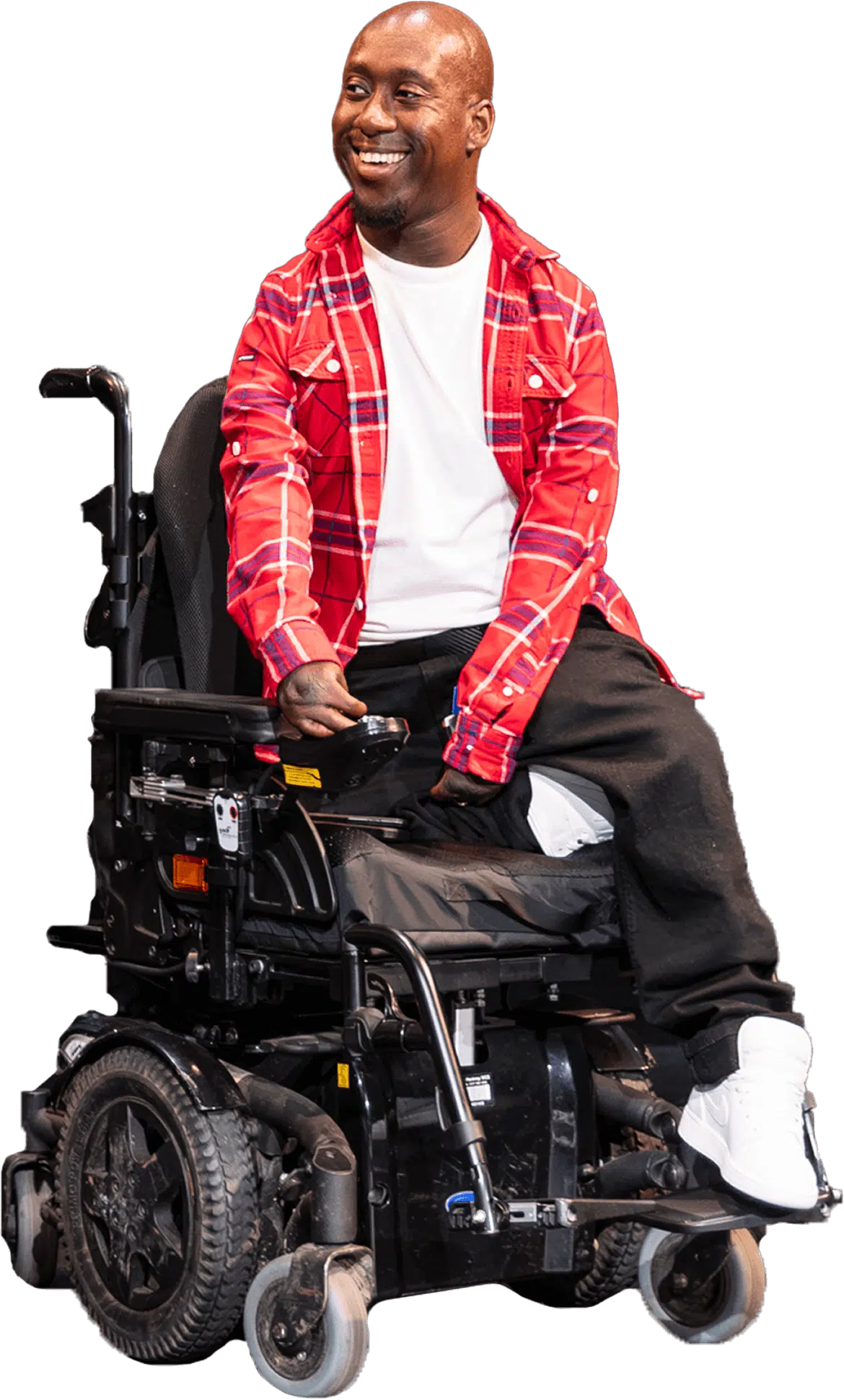 A smiling man wearing a red checked shirt, white t-shirt, black trousers, and white trainers sits in a motorised wheelchair, looking off to the side.