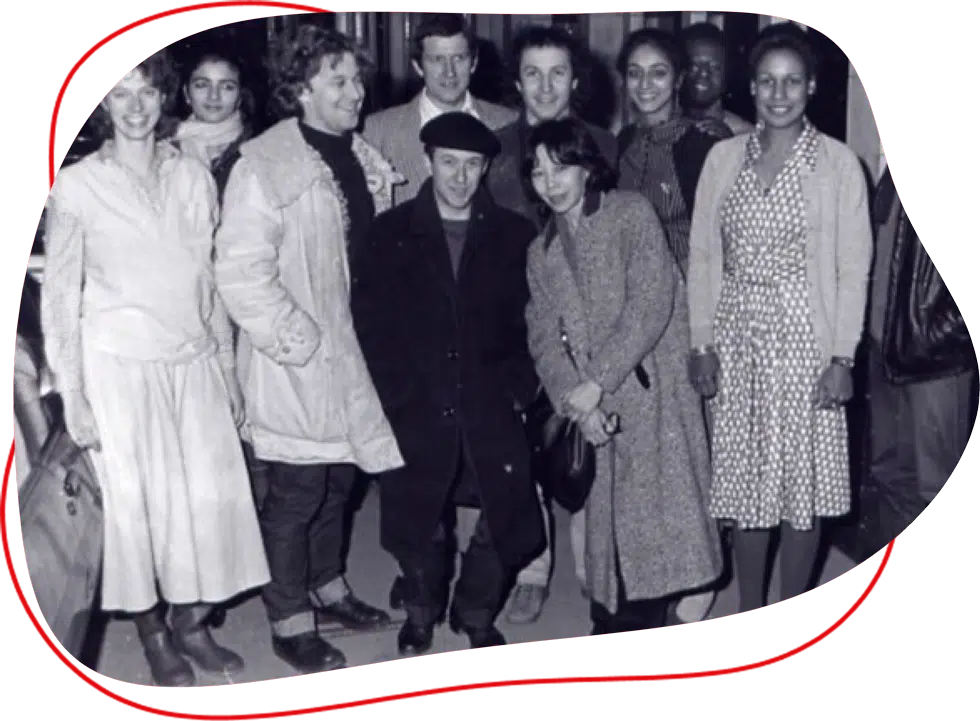 A black-and-white photo of a group of ten adults, men and women, standing closely together and smiling at the camera. Most are wearing coats and winter clothing, suggesting a cold day.