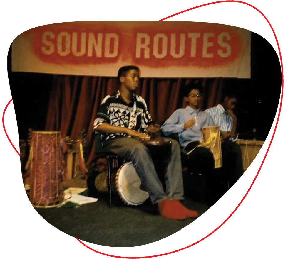 Three people sit on stage playing drums in front of a sign that reads SOUND ROUTES. The person in the centre wears a patterned shirt and red socks, while the others hold different percussion instruments.