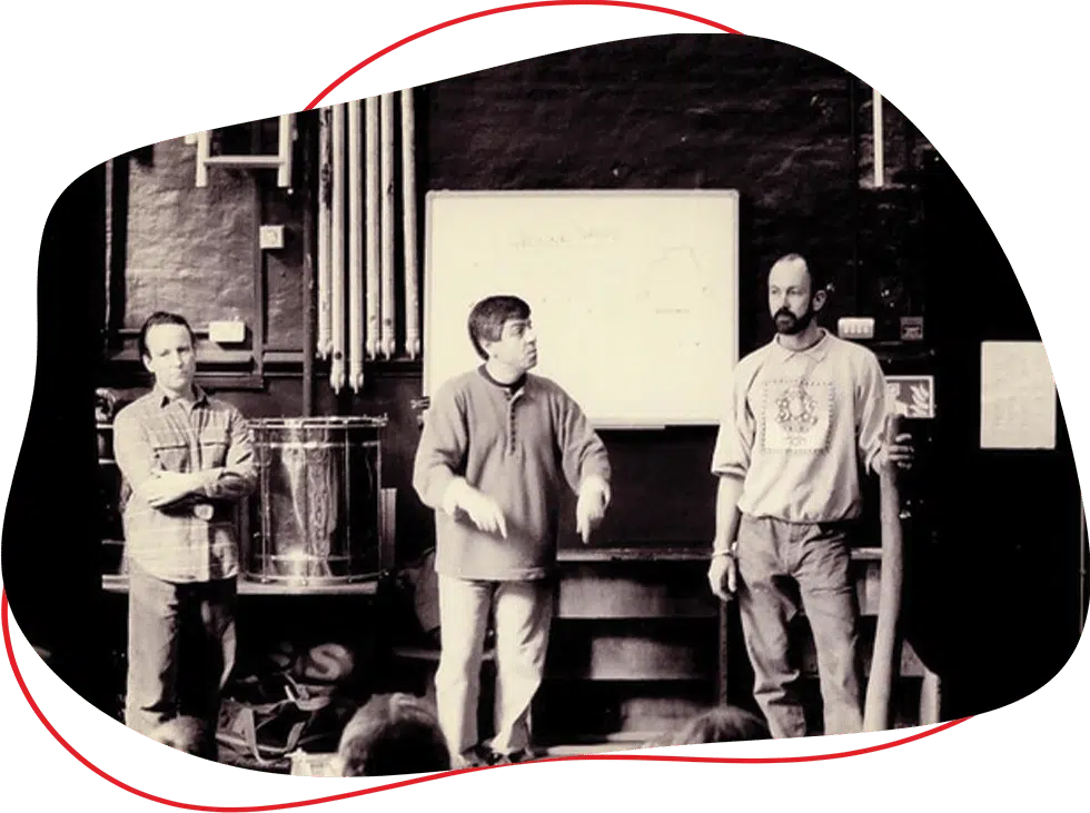 Three men stand in front of a whiteboard in a classroom or workshop setting. One is speaking with his hands raised, whilst the others listen. Pipes, drums, and various equipment are visible in the background.