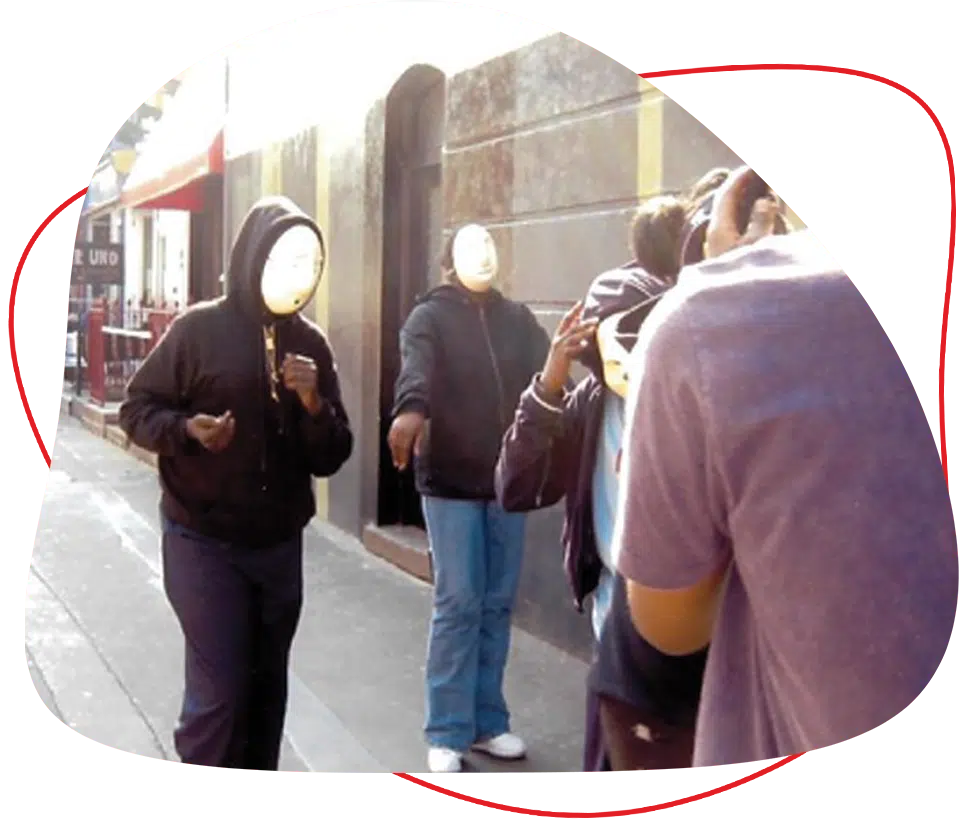 Four people stand on a pavement wearing hooded jackets and white face masks. The background shows a street with buildings and sunlight casting shadows. The atmosphere appears casual.