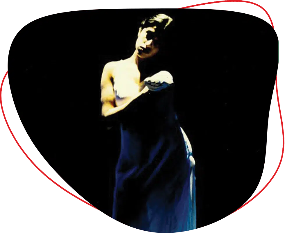 A person in a strapless dress stands in dramatic lighting against a black background, with their head tilted and arms folded, creating a moody, artistic atmosphere.