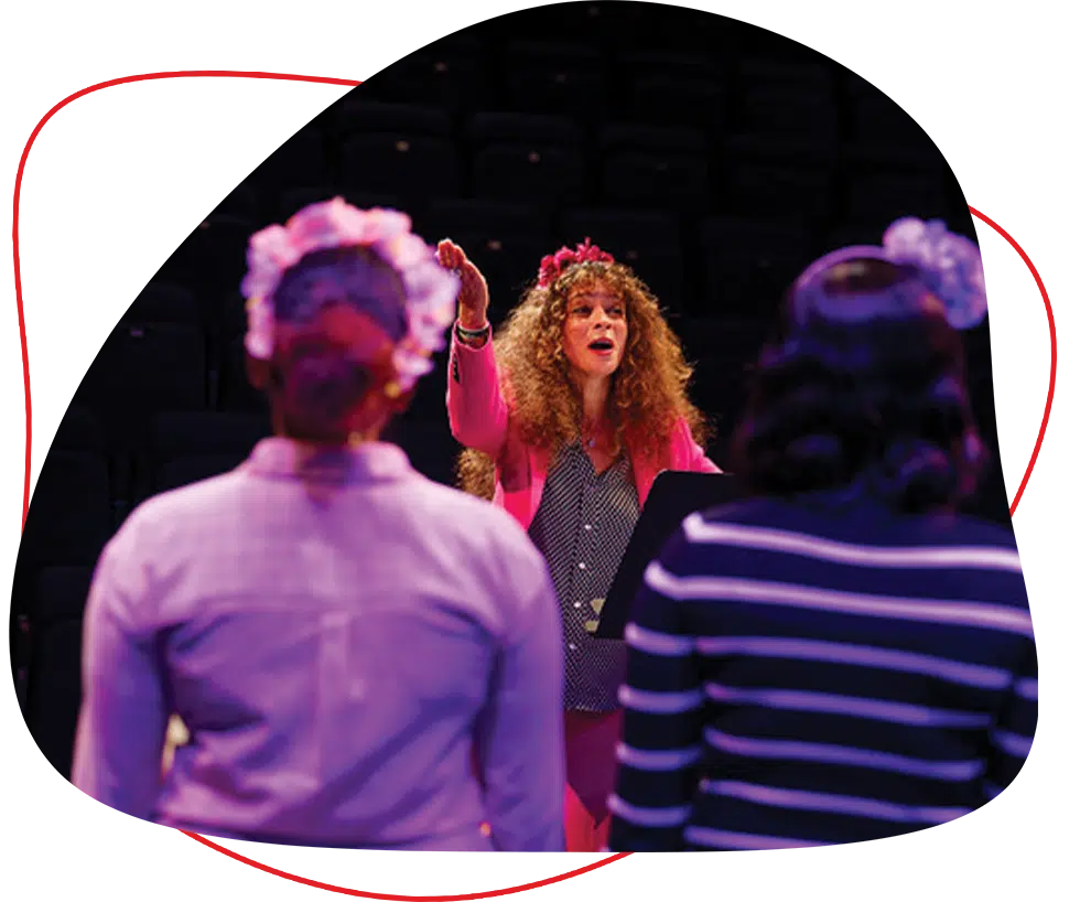 A woman with curly hair and a flower crown conducts two people on stage, both wearing flower crowns, as they face her with their backs to the camera. Dark seats are visible in the background.