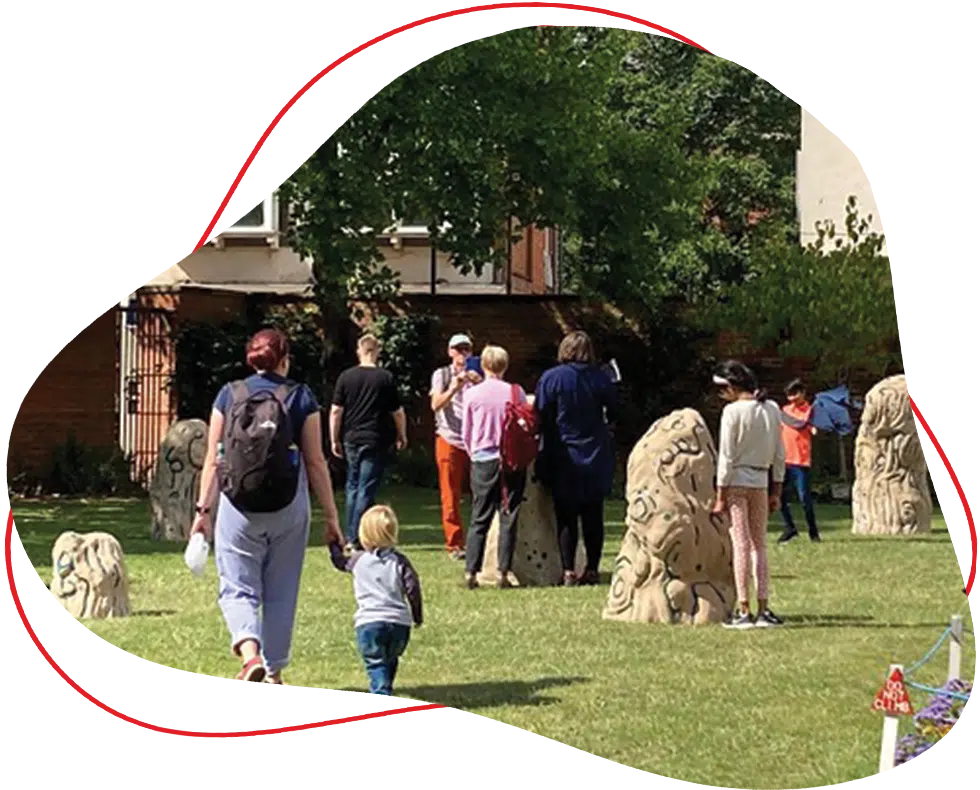 A group of people, including adults and children, walk among sculpted stone pillars on a grassy area with trees and buildings in the background. Some are observing the sculptures, whilst others walk together.