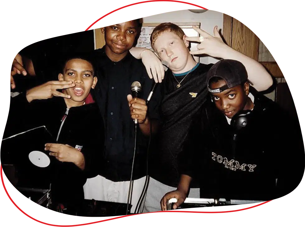 Four teenage boys pose together indoors. One holds a vinyl record, another holds a microphone, and DJ equipment is visible. They smile and gesture playfully, creating a friendly, energetic atmosphere.