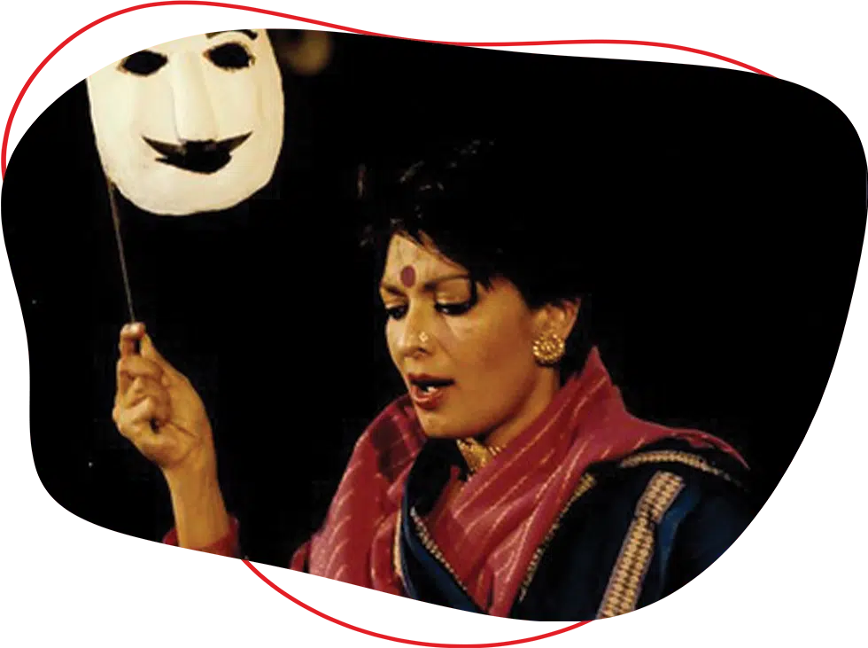 A woman in traditional Indian attire holds a white theatrical mask on a stick. She has short hair, wears a bindi, gold earrings, and is draped in a pink and blue sari, set against a dark background.