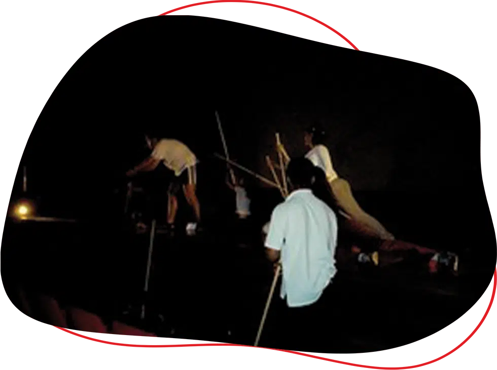 Several people perform on a dark stage, some holding sticks or poles, with one person kneeling in the foreground and others extending their arms or leaning forwards. The scene is dimly lit with a spotlight.