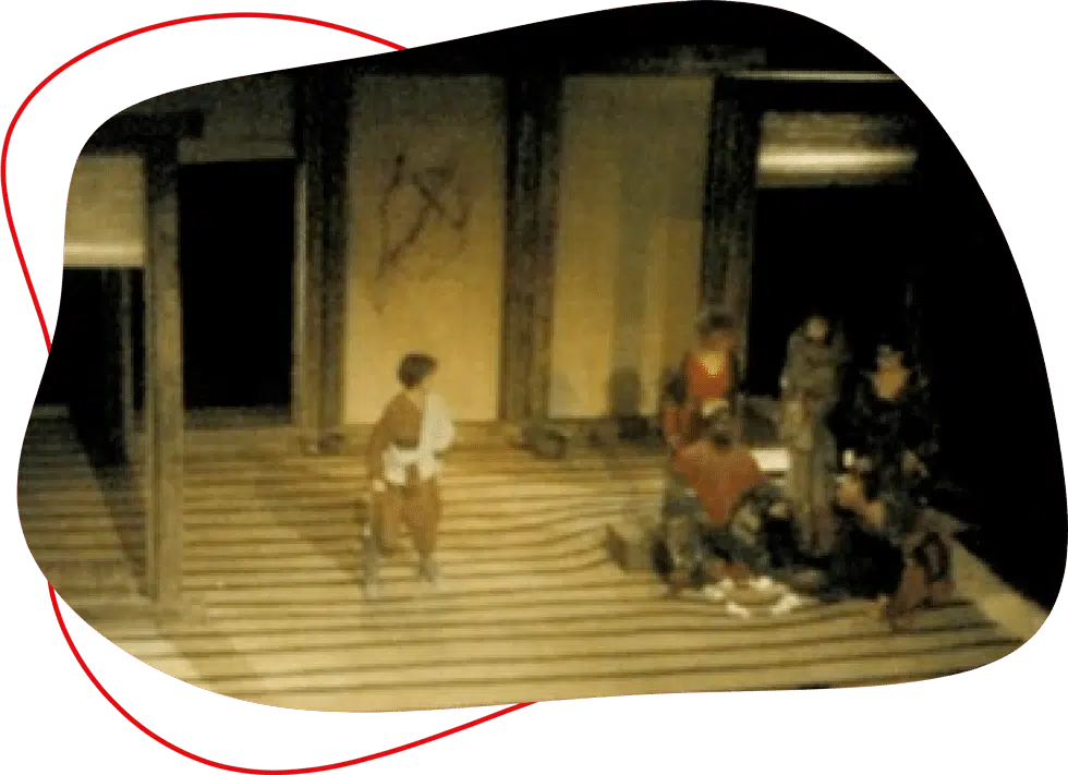 A group of people in costume perform on a dimly lit wooden stage with simple set pieces, including doorways and a wall. Some stand while others sit, suggesting a dramatic scene in a theatre production.