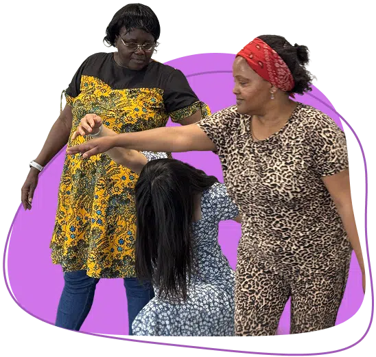 Three women interact in front of a purple abstract shape; one in a yellow patterned dress stands, another in leopard print reaches out, and a seated woman in a blue patterned dress leans forward, her hair covering her face.