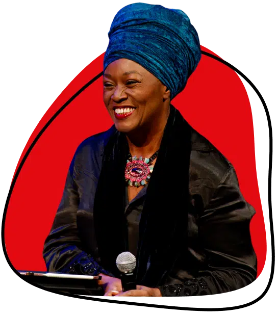 A smiling woman in a blue headwrap and black outfit holds a microphone. She has a bright necklace and stands against a red and white abstract background.