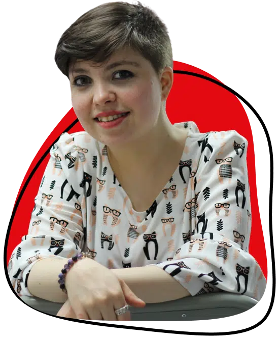 A person with short brown hair smiles whilst leaning on a table, wearing a white shirt patterned with cartoon owls and glasses. The background features a red shape outlined in black.