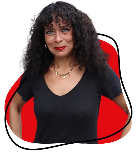 A woman with curly dark hair and blue eyes, wearing a black V-neck top and a gold necklace, stands smiling with her hands on her hips against a white background with red and black abstract shapes.