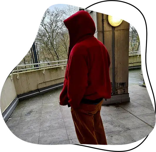 A person wearing a red hooded jacket and brown trousers stands on a balcony, facing away, with bare trees and a cloudy sky in the background.