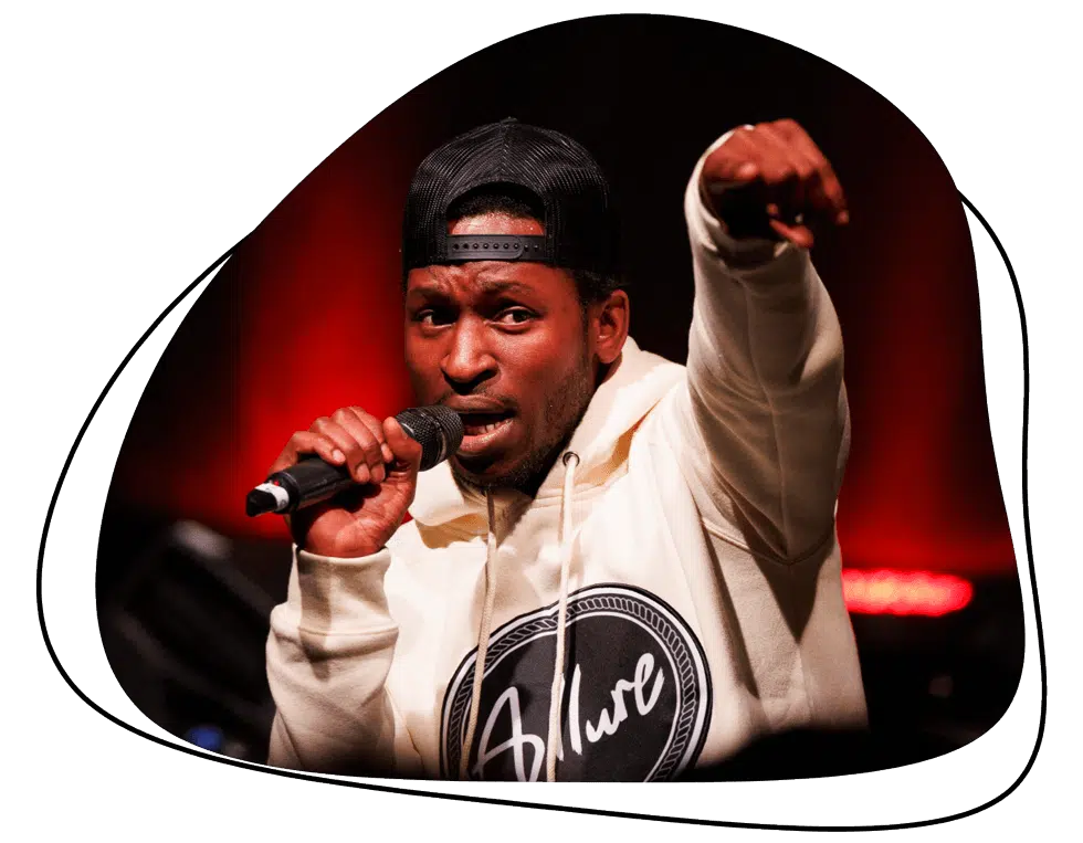 A man wearing a black cap and a white hoodie holds a microphone and points forward whilst performing on stage, with red lights in the background.