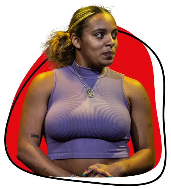 A woman wearing a sleeveless, ribbed lavender crop top and layered necklaces stands with her hands clasped, looking to the side. She has curly hair tied back and a red abstract shape in the background.