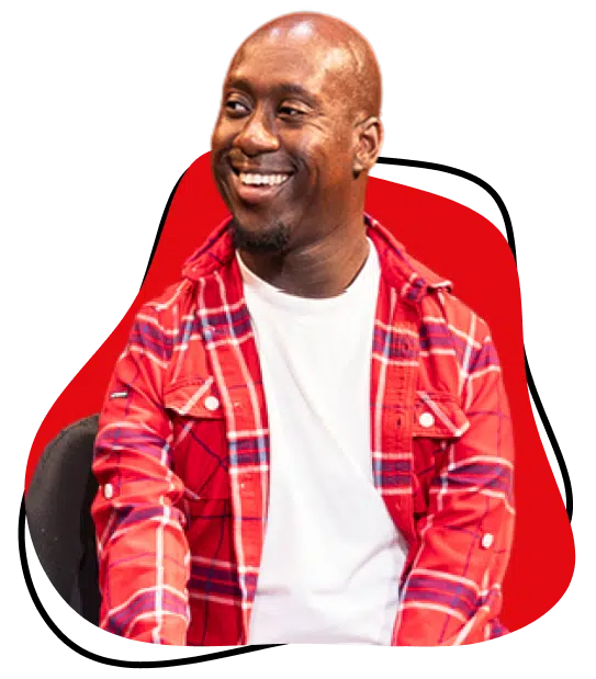 A smiling man wearing a red checked shirt over a white T-shirt sits against a bright red abstract background shape.
