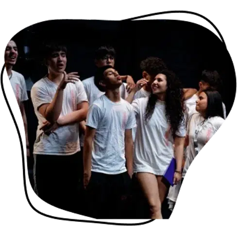 A group of young people wearing white shirts stand together, chatting and smiling against a dark background. One person holds a blue folder, and everyone appears relaxed and happy.