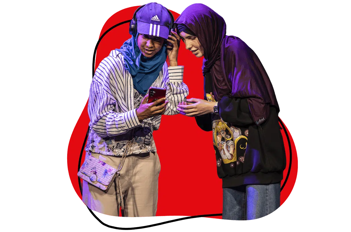 Two young women in headscarves smile while looking at a mobile together. One wears headphones, a cap, and a striped shirt; the other wears a hoodie with an anime design. A red abstract shape is in the background.