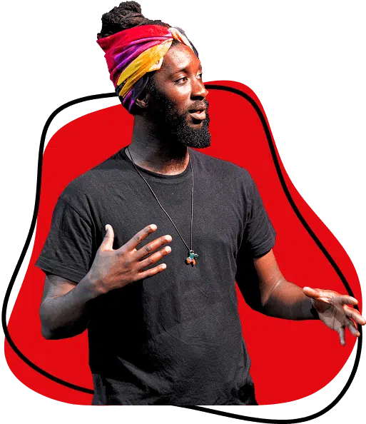 A bearded person wearing a colourful head wrap, black t-shirt, and necklace gestures with their hands whilst speaking, standing in front of a red abstract background.
