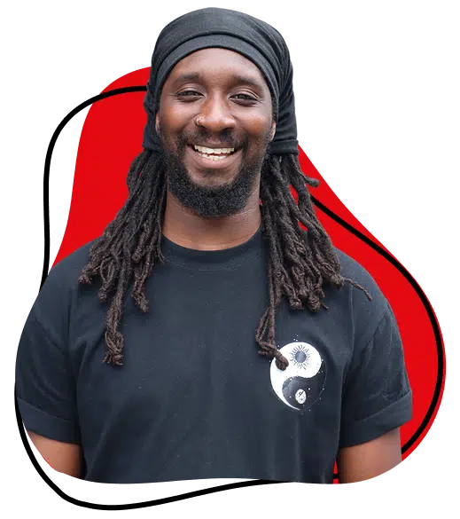 A smiling man with long dreadlocks wears a black headwrap and a black T-shirt featuring a yin-yang design. The background includes a red abstract shape with a black outline.