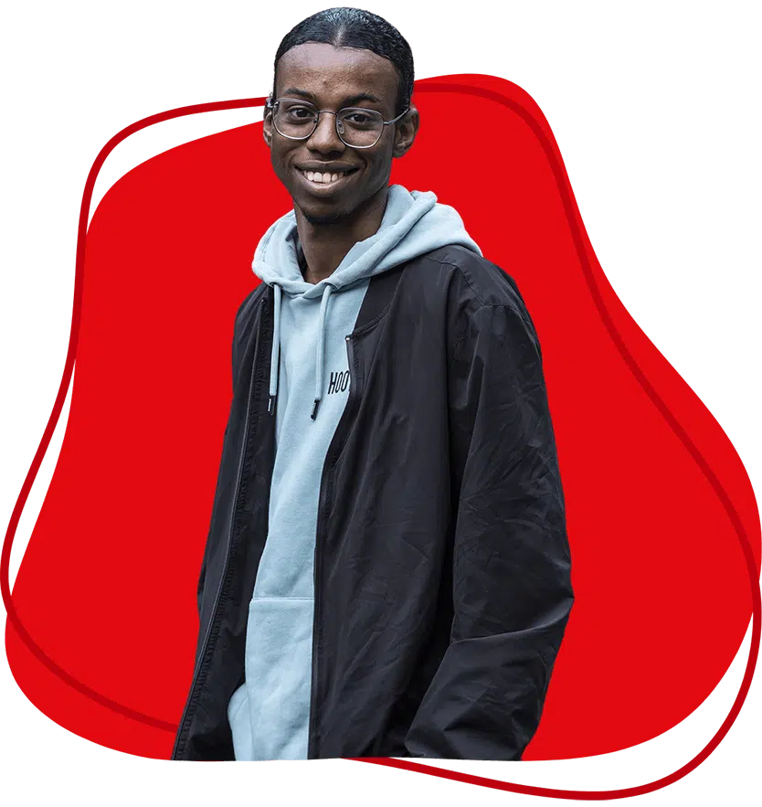 A young man wearing glasses, a light blue hoodie, and a black jacket smiles whilst standing in front of a red abstract background.
