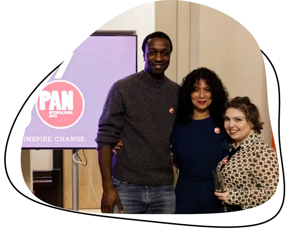 Three people stand together smiling at an indoor event. Behind them, a screen displays a logo with the words PAN INTERCULTURAL ARTS and the phrase INSPIRE. CHANGE.