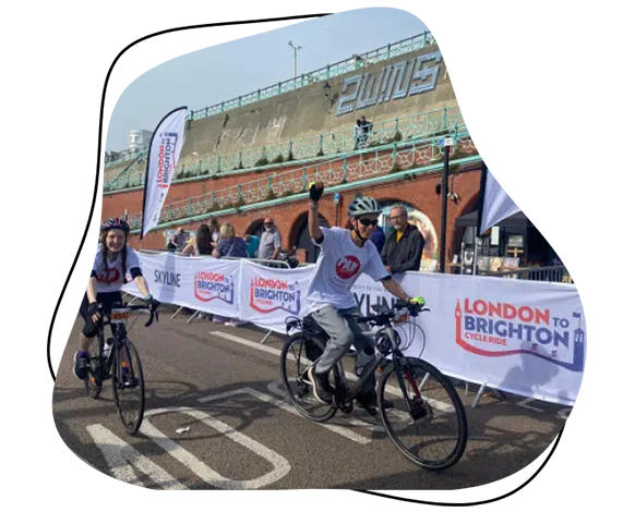 Two cyclists smile and wave as they approach the finish line of the London to Brighton Cycle Ride, with event banners and spectators visible in the background.