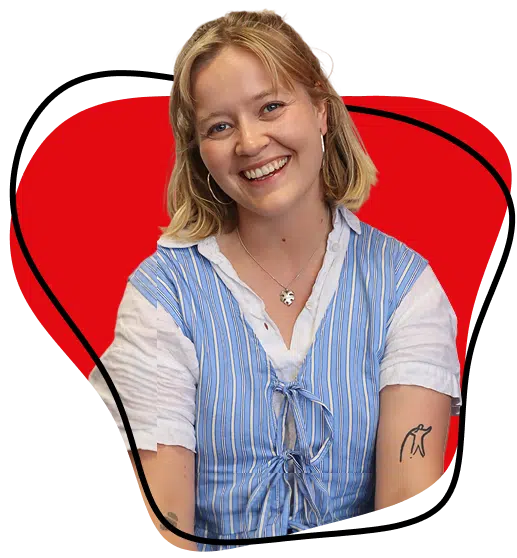 A smiling woman with blonde hair wears a blue and white striped waistcoat over a white shirt. She has hoop earrings, a necklace, and a penguin tattoo on her arm. The background features a red abstract shape outlined in black.