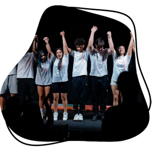 A group of six people on stage, standing in a line with their hands raised together, celebrating or acknowledging an audience in front of them. They are wearing casual clothes and smiling against a dark background.