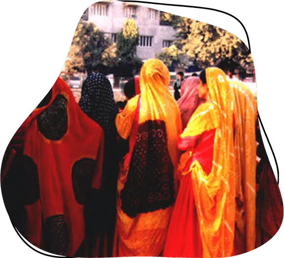A group of women dressed in colourful traditional Indian sarees stand outdoors with their backs to the camera, facing a building and trees in the background.