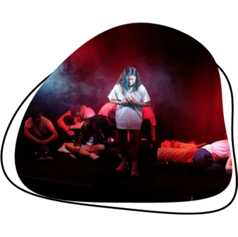 A girl stands alone in the spotlight on stage, looking down, whilst several people sit or lie down behind her amid dramatic red and blue stage lighting and smoke effects.