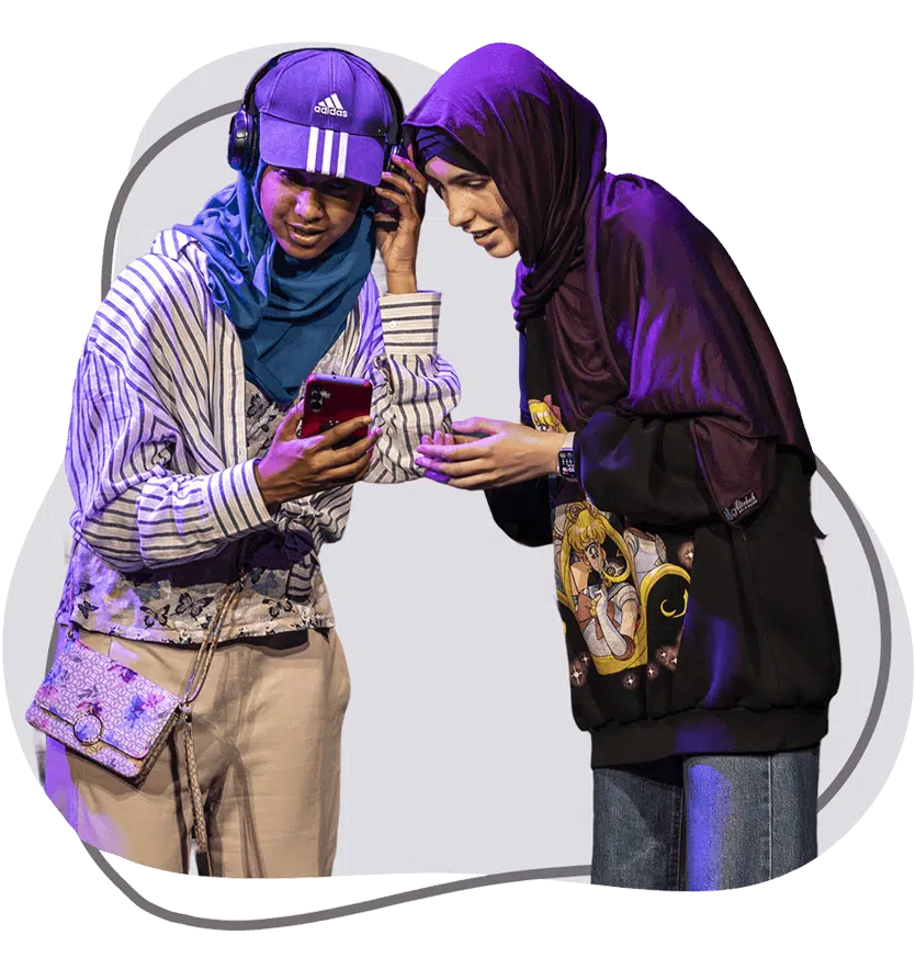 Two young people wearing headscarves and casual clothes look at a smartphone together, smiling. One wears headphones and a cap, while the other wears a hoodie with an anime print.