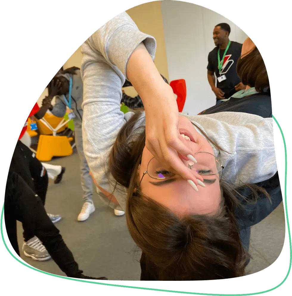 A young woman, wearing glasses and a grey hoodie, playfully poses upside down, holding her face with one hand. Other people in casual clothing are visible in the background, smiling and engaging in activities.