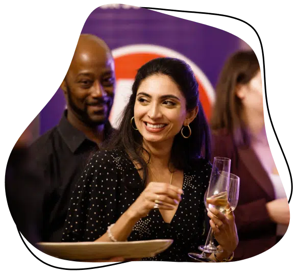 A woman in a black dress with white spots smiles whilst holding a champagne glass at a social event. A man and other people stand behind her, and the background is softly blurred with purple tones.