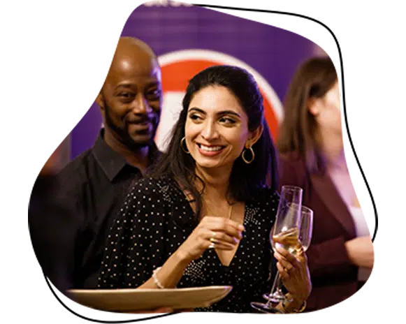 A woman holding a glass of champagne smiles whilst talking at a social event, with a man standing beside her and other people in the background.